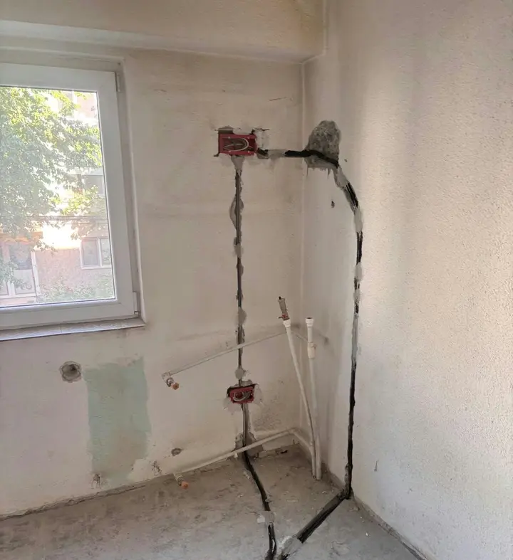 Cabluri în tuburi pe perete și pe pardoseală, cu două doze roșii, într-o încăpere neterminată